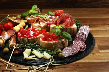Traditional italian antipasto bruschetta appetizer with cherry tomatoes, cream cheese, basil leaves and balsamic vinegar on cutting board with prosciutto, salami, cheese