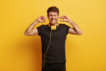 Turn volume, please! Annoyed young man pluggs ears and clenches teeth, dissatisfied with loud rock...