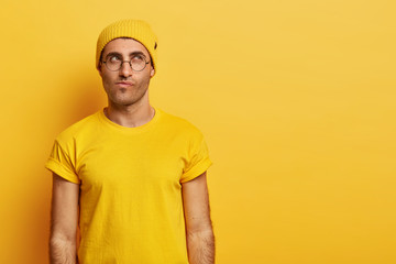 Contemplative Caucasian guy looks with thoughtful expression upwards, focused above, purses lips, tries to make decision, wears round eyewear, bright yellow hat and t shirt, blank space aside