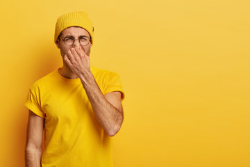 Disgusted young hipster pinches nose with fingers, looks in disgust as smells something stink, wears spectacles, yellow hat, t shirt, stands in stuido with blank copy space. Fie, unpleasant smell - obrazy, fototapety, plakaty