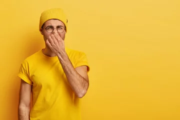 Foto op Canvas Disgusted young hipster pinches nose with fingers, looks in disgust as smells something stink, wears spectacles, yellow hat, t shirt, stands in stuido with blank copy space. Fie, unpleasant smell © Wayhome Studio