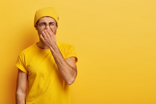 Disgusted Young Hipster Pinches Nose With Fingers, Looks In Disgust As Smells Something Stink, Wears Spectacles, Yellow Hat, T Shirt, Stands In Stuido With Blank Copy Space. Fie, Unpleasant Smell