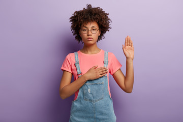 Confident serious woman with curly hair makes sincere promise or oath, keeps one hand on heart, solemnly swears, raises palm, demonstrates loyalty gesture being honest poses against purple background. - obrazy, fototapety, plakaty