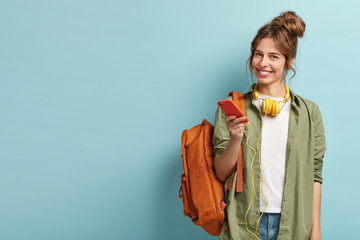 Studio shot of glad female model holds modern smartphone gadget, reads publication and listens...
