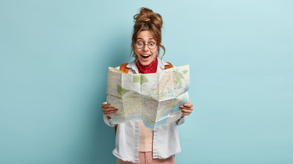 Photo of happy shocked female stares at map, enjoys adventure during summer vacations, joyful to...