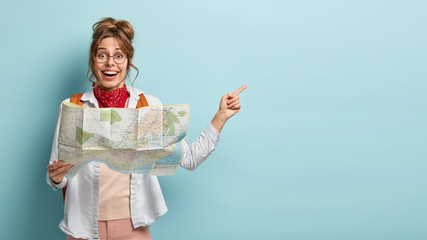 Smiling optimistic young European female guide holds map, shows direction to tourist away, demonstrates destinations of showplace, dressed casually, isolated on blue wall, searches right route - obrazy, fototapety, plakaty