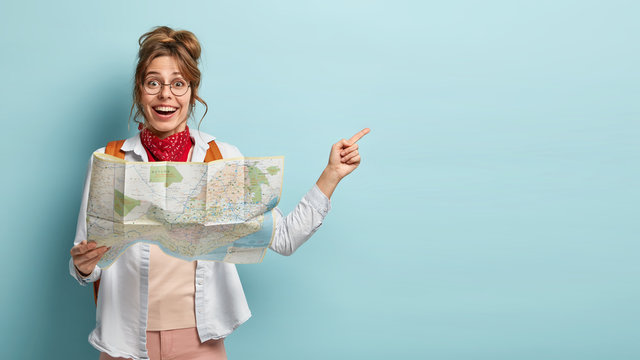 Smiling optimistic young European female guide holds map, shows direction to tourist away, demonstrates destinations of showplace, dressed casually, isolated on blue wall, searches right route