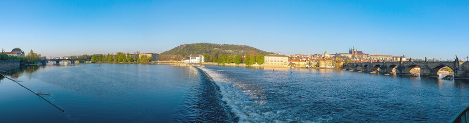 Outdoor sunny panoramic scenery of Vltava river with riverside, weir on the river, famous Charles bridge, and city skyline with background of Praha Castle on the hill in Prague, Czech Republic. 