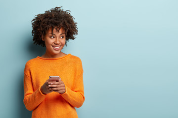 Photo of cheerful delighted African American woman types sms on modern cell phone device, enjoys...