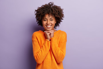 Horizontal shot of happy Afro American woman with toothy smile, keeps both hands under chin, expresses positiveness as listens pleasant words of praise, has nice day isolated over purple background.