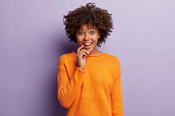 Studio shot of glad mirthful woman keeps fore finger on lips, smiles delighted and upbeat at camera, wears orange jumper, shows white teeth, isolated over purple background. Positive emotions