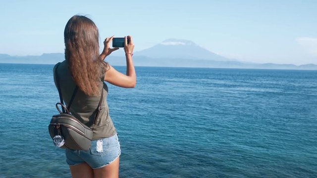 Back side view of girl traveler with backpack takes photo of ocean and volcano on phone