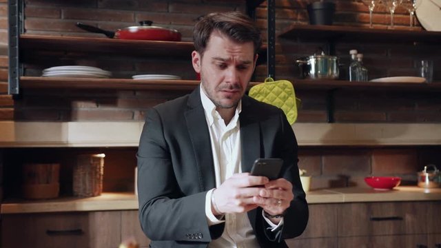 Handsome emotional Caucasian man in formal suit sitting at the kitchen table and actively messaging via smartphone, reacting negatively on received sms. Bad mood, bad news, messaging app