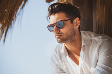 Young and handsome man with sunglasses looking. Warm summer lights, he wears a white shirt. Light short beard. Fashionable hair.