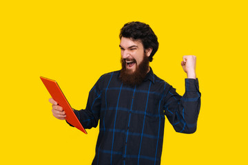 Photo of handsome man with beard is screaming and celebrating with rised arm, while holding a tablet, over yellow wall