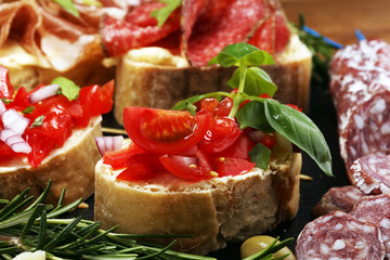 Traditional italian antipasto bruschetta appetizer with cherry tomatoes, cream cheese, basil leaves and balsamic vinegar on cutting board with prosciutto, salami, cheese