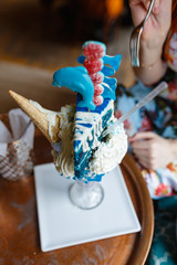 Fototapeta premium Close-up of crazy giant ice cream in large glass with waffle cone, piece of cake, marmalade, cream, and milk cocktail inside. Insanely delicious and high-calorie sweet ice cream milkshake in cozy cafe