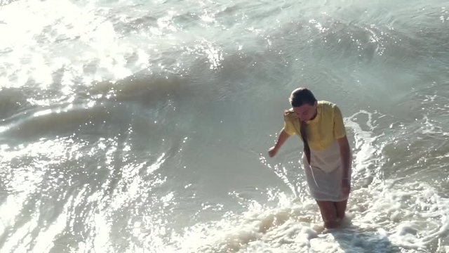 A beautiful, sad girl in a yellow-white dress and long blond, wet hair comes out of the muddy, muddy sea. She is hit by waves with foam. Bright sun, glare on the water.