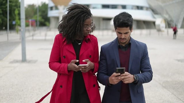 Pretty Afro-american woman in spectacles and rose coat and handsome mixed-race man in navy blue suit walking along street, man showing photos on phone, they talking. Communication, lifestyle concept