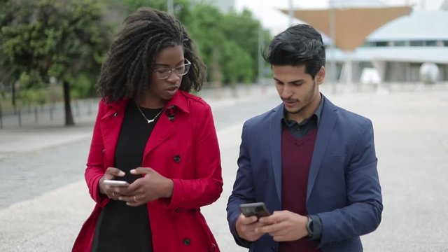 Pretty Afro-american woman in spectacles and rose coat and handsome mixed-race man in navy blue suit walking along street, man showing photos on phone. Dolly shot. Communication, lifestyle concept
