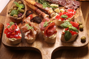 Traditional italian antipasto bruschetta appetizer with cherry tomatoes, cream cheese, basil leaves and balsamic vinegar on cutting board with prosciutto, salami, cheese