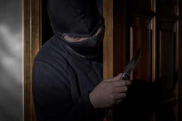 Thief with a knife in his hand and a hat with a slit for the eyes sneaks into the house through the...