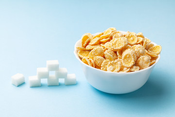 High sugar in flakes. Pile of sugar cubes and plate with cereal on blue background.