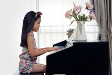Side view of young little Asian cute girl playing electronic piano at home. Seen from side view while she pressing piano keys by both hands. She practicing music skill. Hobby at home concept.
