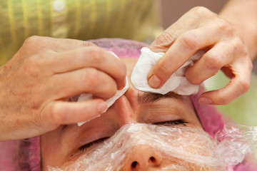 Naklejka premium A young girl is lying on a couch during cosmetic procedures with a mask on the face above which beautician woman squeezes body fat and pimples with wipes. Cleansing and make up.
