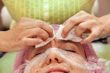 A young girl is lying on a couch during cosmetic procedures with a mask on the face above which beautician woman squeezes body fat and pimples with wipes. Cleansing and make up.