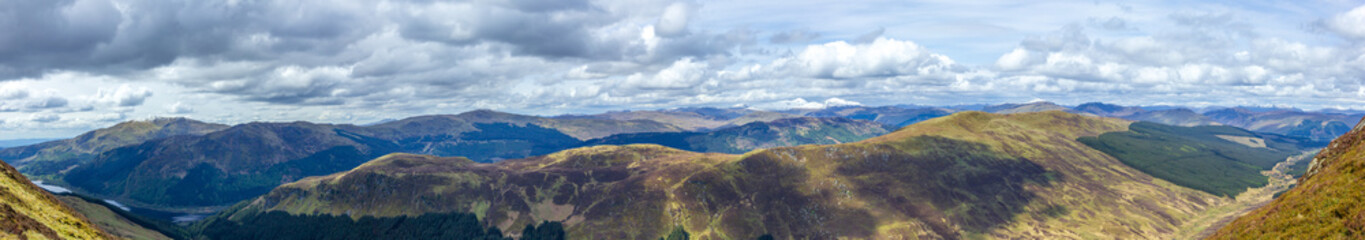 A panorama view of a Scottish mountain range with lakes, pines forest under a majestic cloudy white sky