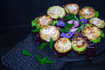 On a black tray are lettuce leaves, and in them are mugs of roasted zucchini. They are watered with sauce. In the middle are the flowers of violets and garlic cloves. 