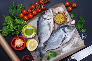 Dorado fish on a brown wooden board on a dark blue background. Next to cherry tomatoes, spinach, pepper, spices for fish, lemon and olive oil