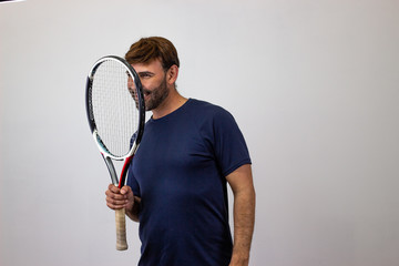 Portrait of handsome young man playing tennis holding a racket with brown hair and a funny expression, facing forwards and looking at the horizon. Isolated on white background.