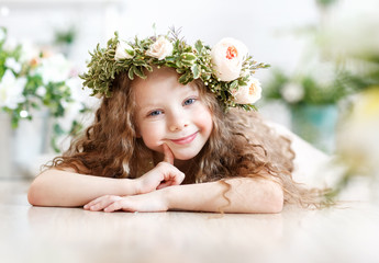 Kid - girl with blond hair in a flower crown. Beautiful little girl in a bright studio lying on the floor and looks into the camera