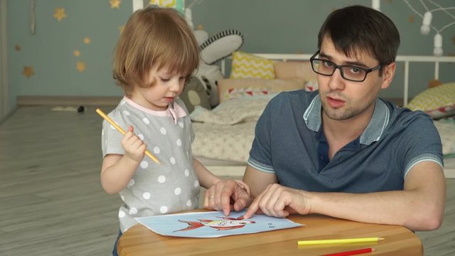 Young attractive father in glasses with little cute girl daughter sit at table in children's room at home and paint goldfish on sheet of paper with colored pencils. Children's creativity. Close-up.
