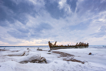 Old broken boat wreck and rocky beach in wintertime. Frozen sea, evening light and icy weather on...
