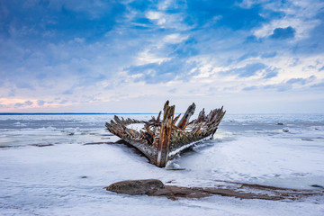 Old broken boat wreck and rocky beach in wintertime. Frozen sea, evening light and icy weather on shore like fairy tale country. Winter on coast. Blue sky, white snow, ice covers the land.
