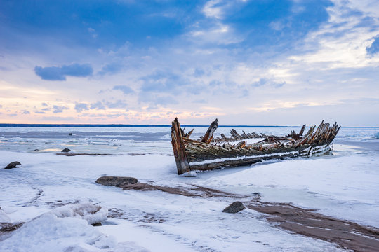 Old broken boat wreck and rocky beach in wintertime. Frozen sea, evening light and icy weather on shore like fairy tale country. Winter on coast. Blue sky, white snow, ice covers the land.