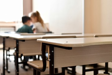 A row of tables, on the last empty briefcase hook in sharpness. On the first table, two children sit side by side and whisper. In the classroom, flooded with light from the window.