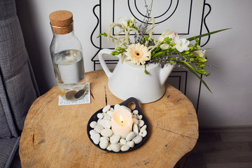 a bowl with a burning candle decorated with white pebbles, glass bottle with water and cork and next to it a teapot with floral decoration, higher position