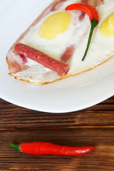 Red chili peppers, small sausages and bacon and eggs are in a white plate on a natural wooden surface of pine boards. Daylight