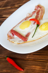 Fried eggs with bacon, red chili pepper and small sausages lie in a white plate on a natural wooden surface of pine boards. Daylight
