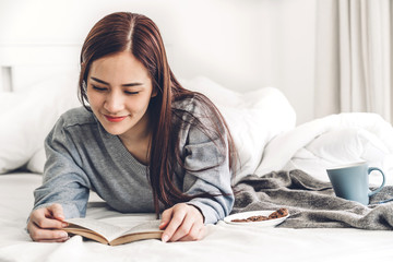 Young woman relaxing reading book and drinking cup of hot coffee or tea  on bed at home