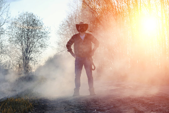 A man is wearing a cowboy hat and a loso in the field. American farmer in a field wearing a jeans hat and with a lasso in the smoke of a fire. A man walks through a burning field