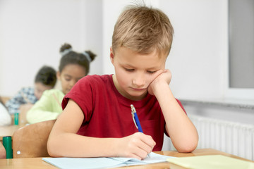 Naklejka na ściany i meble Schoolboy at desk writing with pen in copybook.