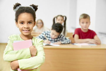 Girl standing in class, holding books, looking at camera.