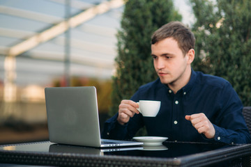 Handsome man drink cappuchino in cafe on the terrace and look into his laptop