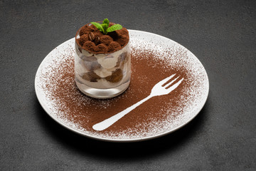 Classic tiramisu dessert in a glass on plate with fork silhouette on dark concrete background