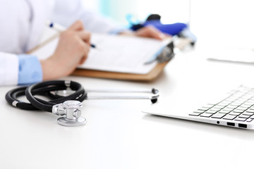 Closeup of stethoscope. Female doctor fills up medical form while sitting at the desk in hospital. Healthcare, workplace and cardiology in medicine concept